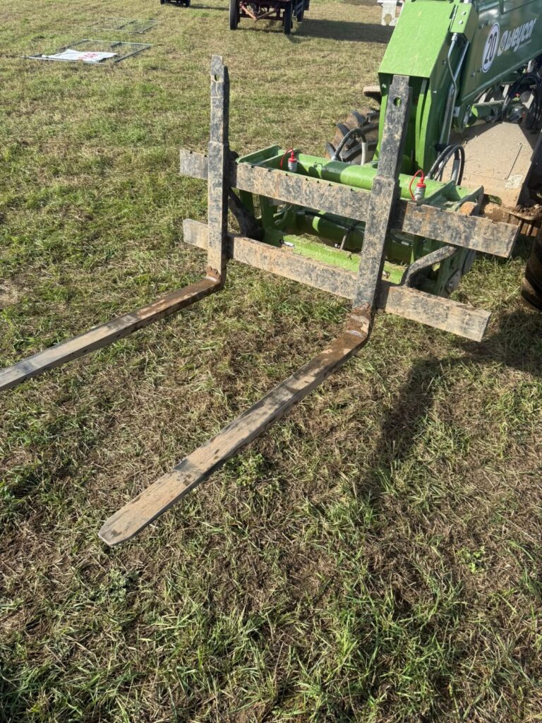 Palettengabel mit zwei Zinken an einem grünen Hoflader, abgestellt auf der Wiese